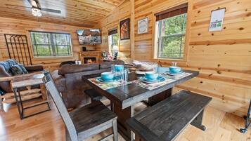 Interior acolhedor de uma cabana com uma mesa de jantar de madeira para quatro pessoas, lareira e uma grande janela com vista para o verde.