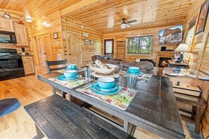 A wooden cabin interior with a dining table set for four, a sofa, a fireplace, and a television mounted on the wall.