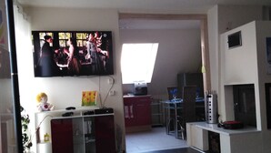 Un salon moderne doté d’un téléviseur à écran plat fixé au mur, d’un coin repas avec une table et des chaises, et d’une cuisine équipée d’un réfrigérateur et d’un four à micro-ondes.