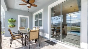 Une terrasse ensoleillée avec une table et des chaises, un ventilateur de plafond et une vue sur une piscine, visible à travers des portes vitrées.