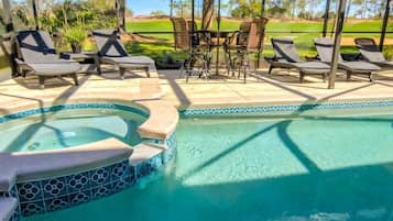 A pool area with lounge chairs and a hot tub, surrounded by a patio with a view of a golf course.