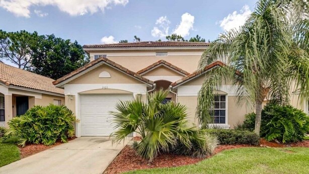 A single-story house with a beige facade, a white garage door, and a small front yard with palm trees and shrubs.