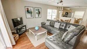 A living room with a grey leather sofa, a coffee table, and a TV on a stand. There are framed pictures on the wall and a kitchen area in the background.