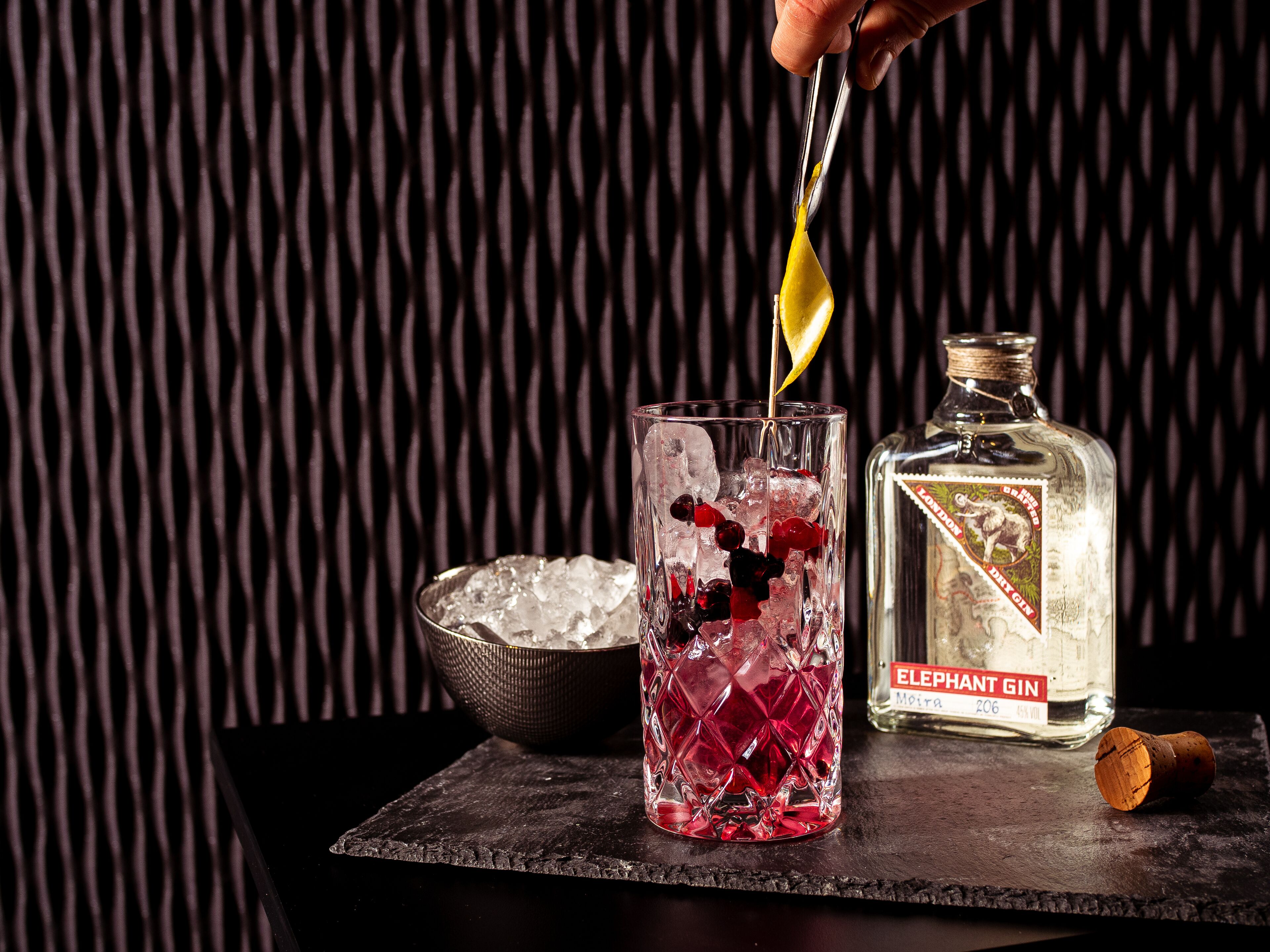 A glass with ice and garnishes, a bottle of Elephant Gin, and a cork on a slate surface.