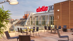 Eine Dachterrasse mit Außenbestuhlung, ein modernes Gebäude mit dem Schriftzug "aletto HIO" und einem klaren Himmel.