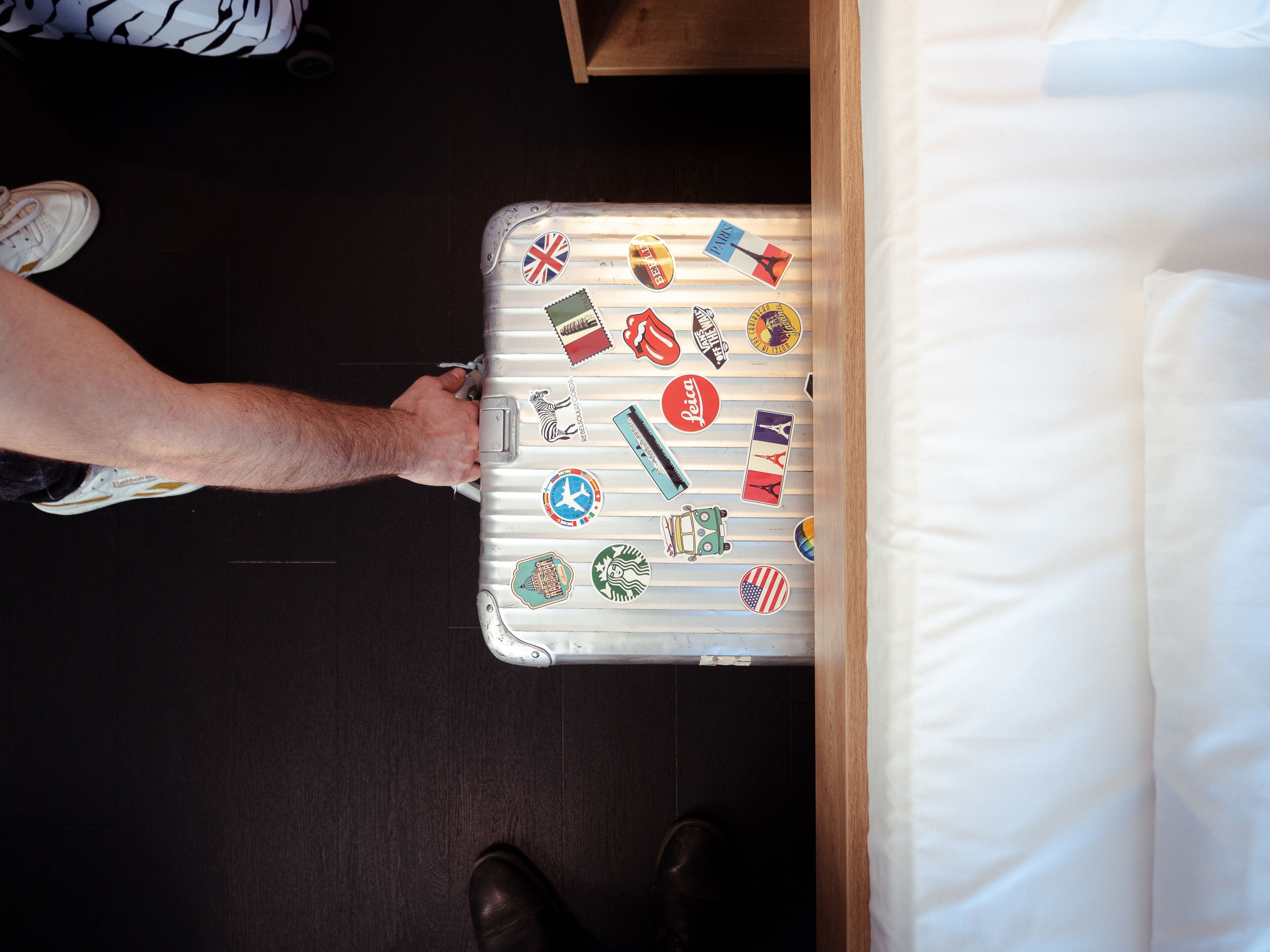 A suitcase with travel stickers, a wooden floor, and a white bed.