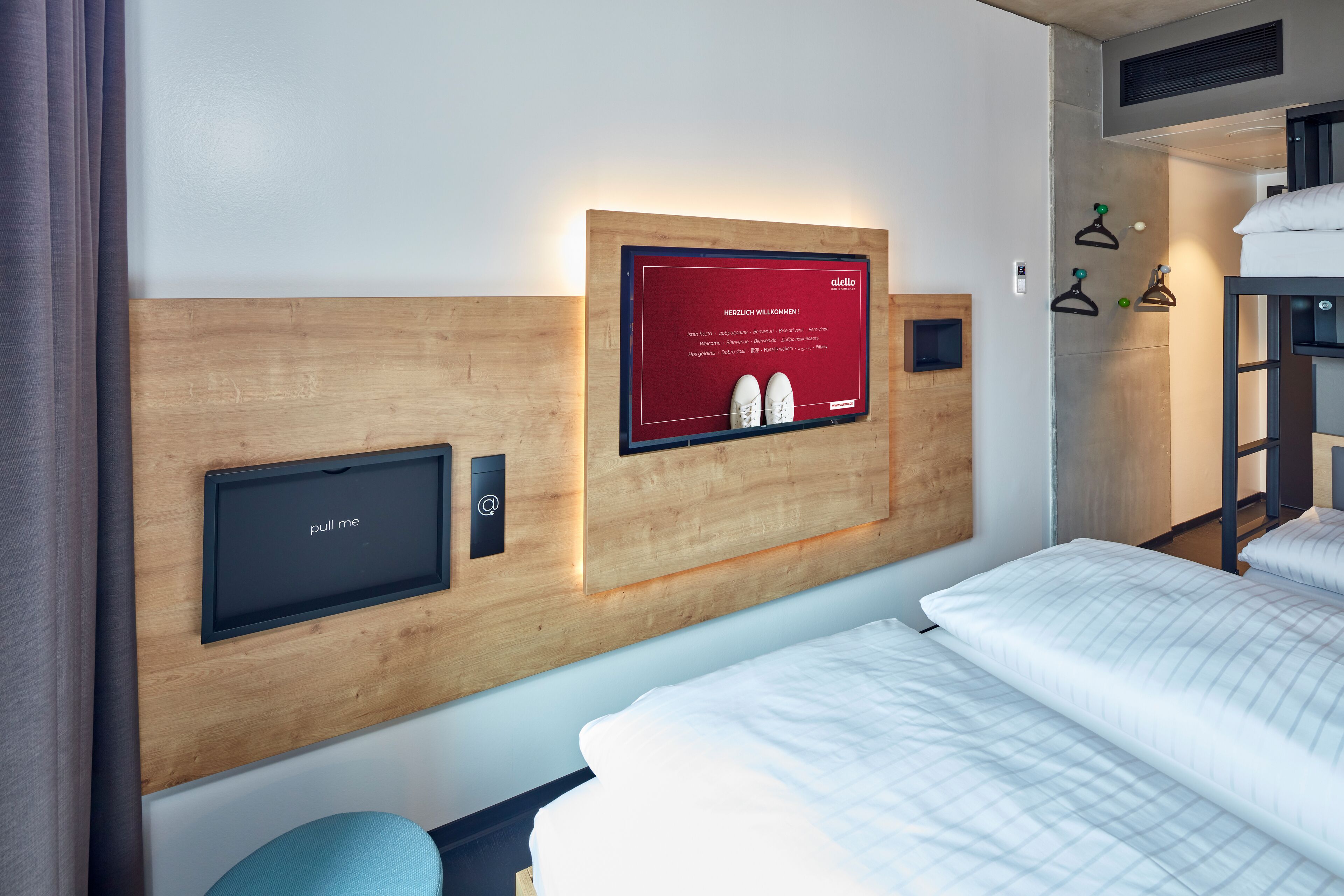 Une chambre d’hôtel avec une tête de lit en bois, un téléviseur fixé au mur, un écran numérique, un lit superposé et un porte-chaussures mural.