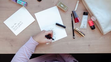 A person writing on a piece of paper with a pen, surrounded by various items like a bottle, a lipstick, and a handbag on a wooden table.