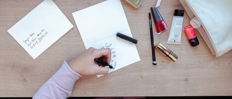 A person writing on a piece of paper with a pen, surrounded by various items like a bottle, a lipstick, and a handbag on a wooden table.