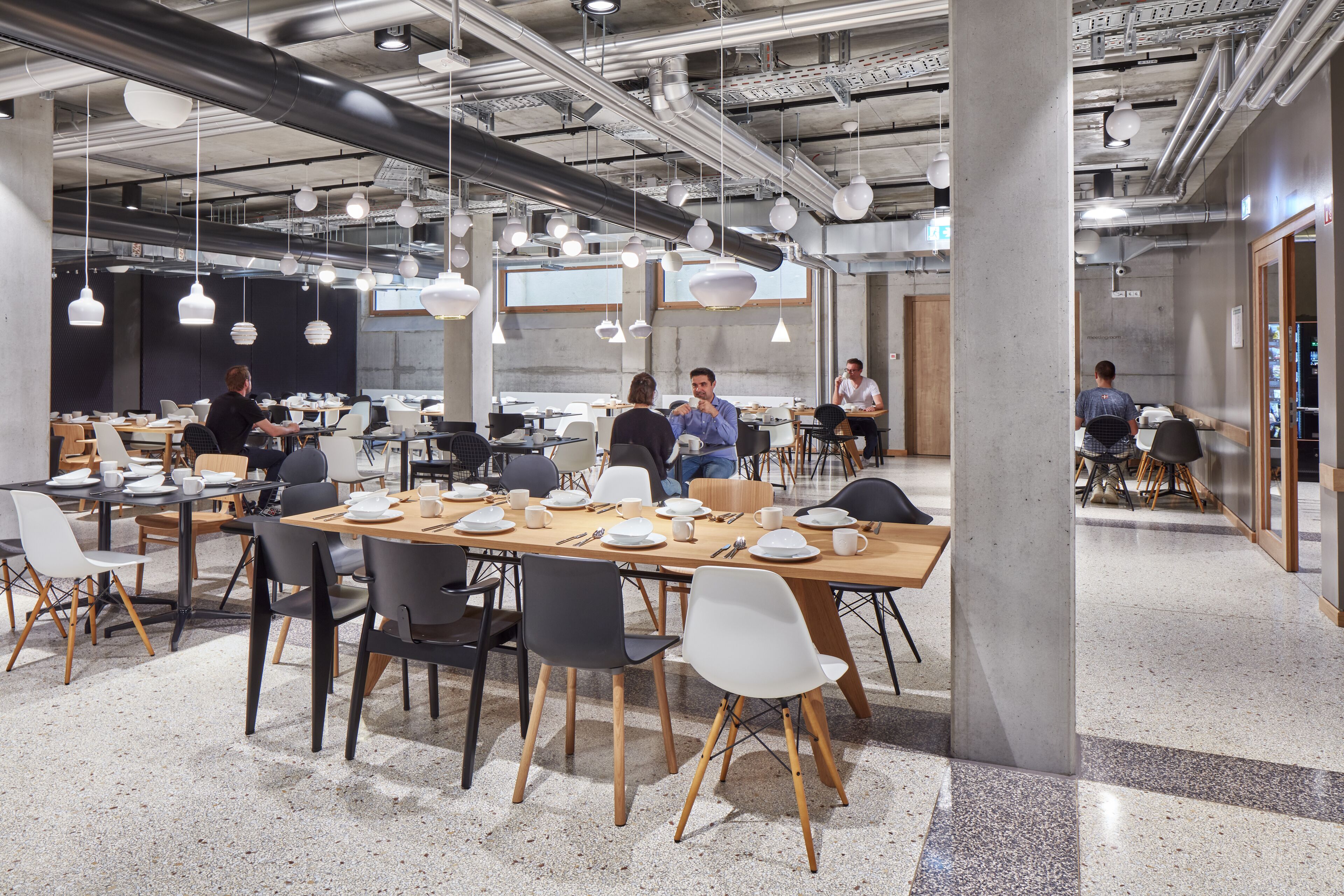 A modern dining area with tables set for meals, hanging pendant lights, and a visible kitchen area in the background.