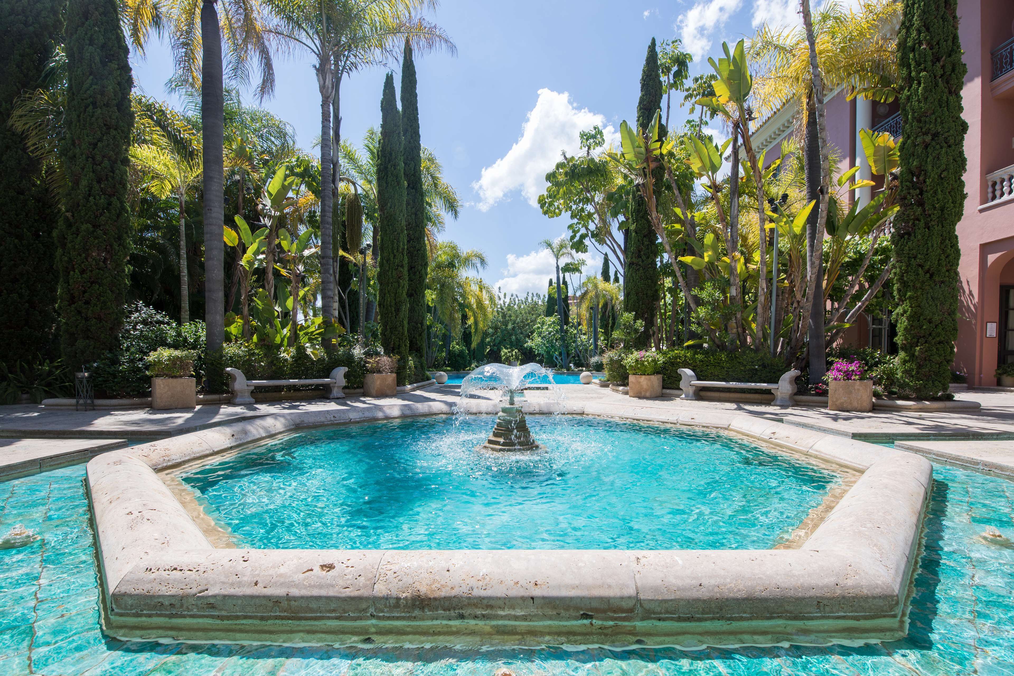 A rectangular pool with a central fountain, surrounded by tall palm trees and cypress trees, with a clear blue sky above.