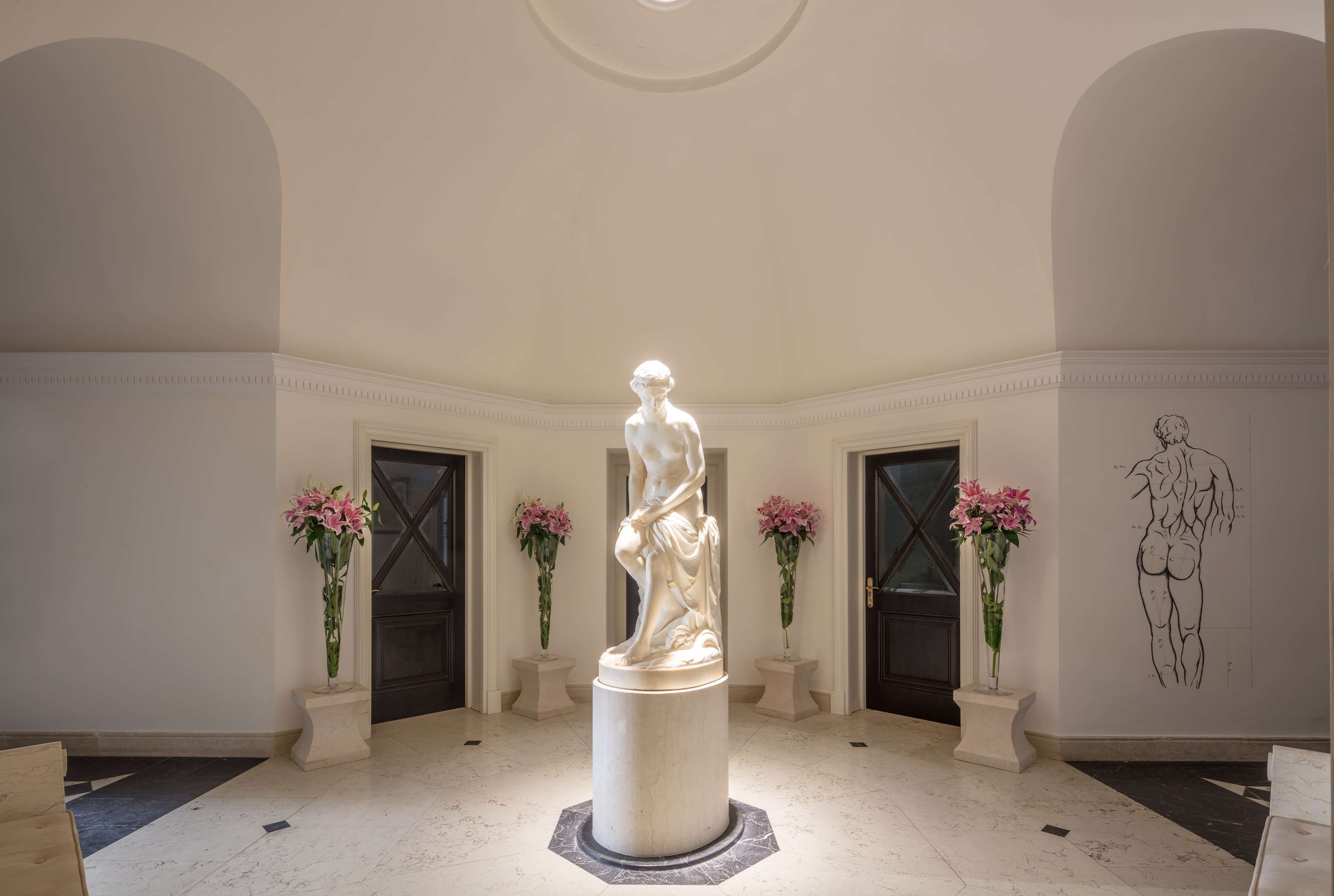 Una estatua de mármol en un vestíbulo con techo alto, dos puertas y arreglos florales.