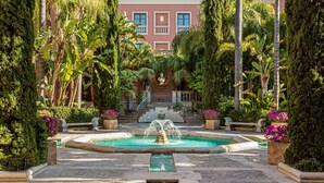 Un patio con una fuente, rodeado de palmeras y plantas en macetas, que conduce a un edificio de fachada roja y varias ventanas.