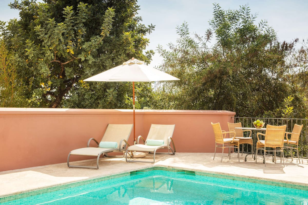 Un espace aménagé au bord de la piscine, avec des chaises longues et une table, entouré d’arbres et bordé d’un mur rose.