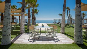 Un área de comedor al aire libre con mesa y sillas, rodeada de palmeras y vista al mar.