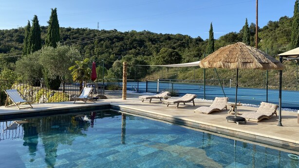 Piscina cristalina com espreguiçadeiras e guarda-sóis, cercada por uma vedação e árvores.