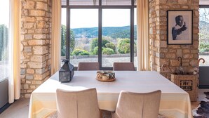 Uma sala de jantar com parede de pedra, grandes janelas e uma mesa com cadeiras.