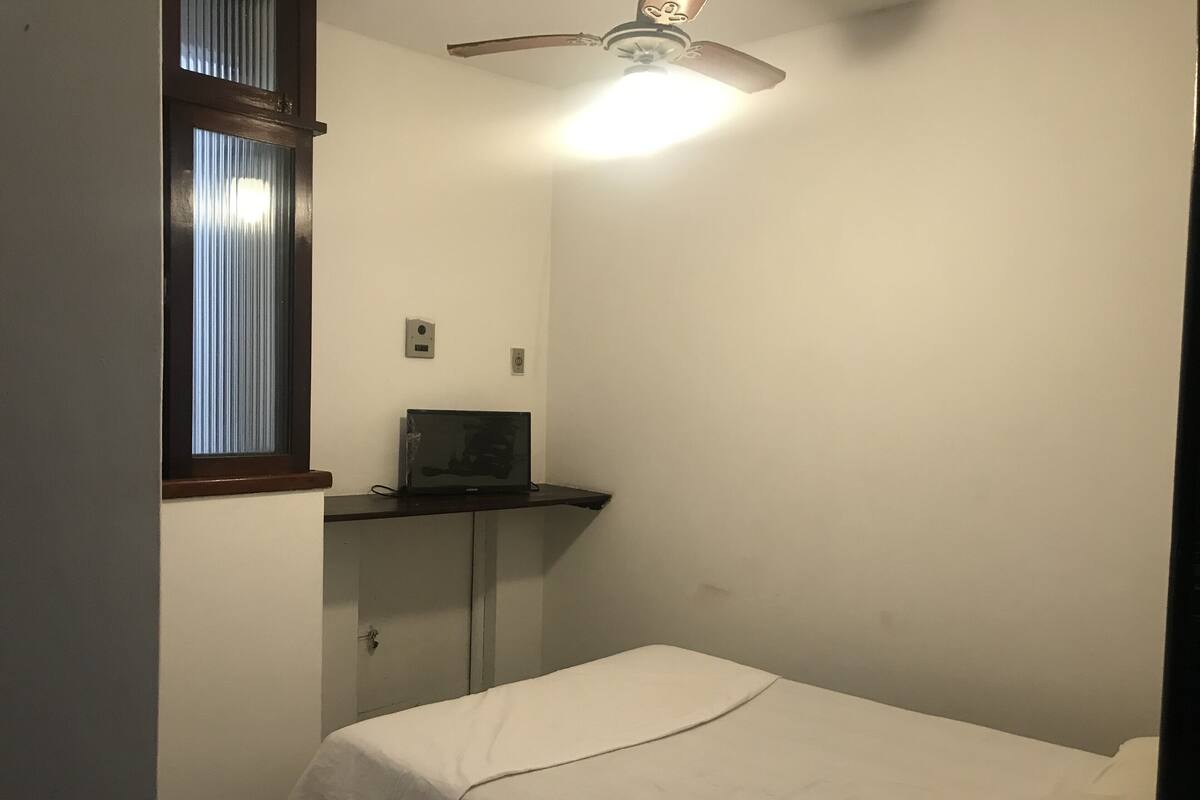 Una habitación pequeña y poco amueblada con una cama individual, un escritorio con una computadora portátil, una ventana con persianas y un ventilador de techo.