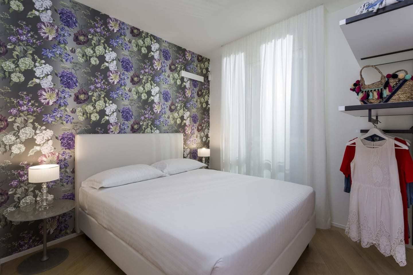 Camera d'albergo con un letto grande, carta da parati floreale, un comodino con una lampada e un appendiabiti con un vestito.