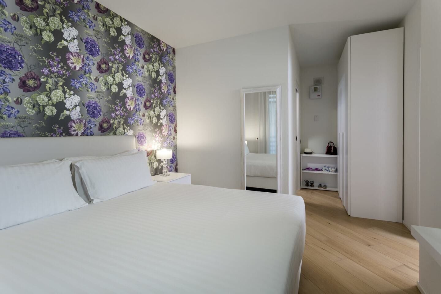 Un dormitorio con una cama grande, una mesita de noche con lámpara, papel tapiz floral y un vestidor.
