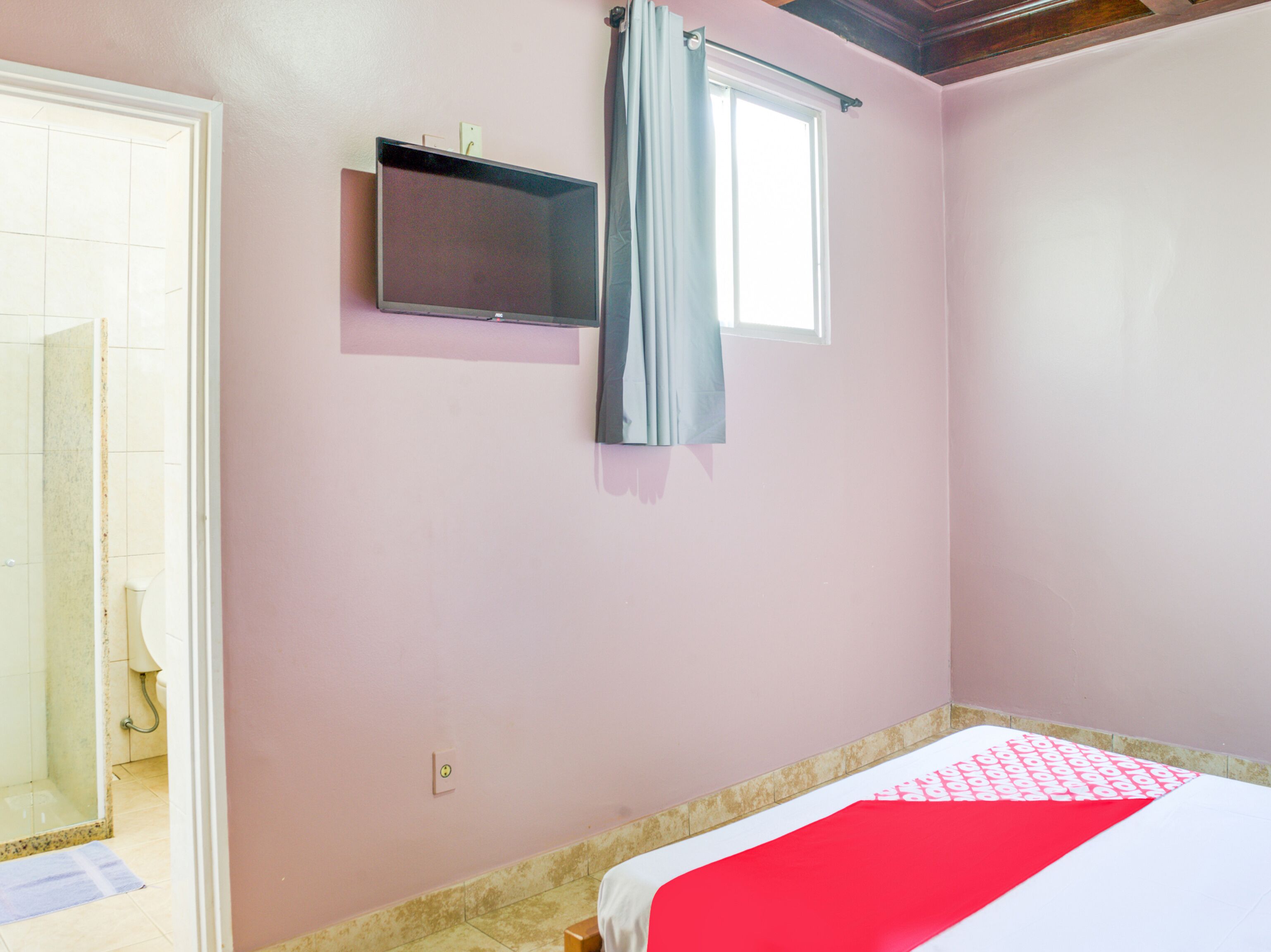 Un dormitorio con cama, televisor montado en la pared, ventana con cortinas y baño a la vista al fondo.