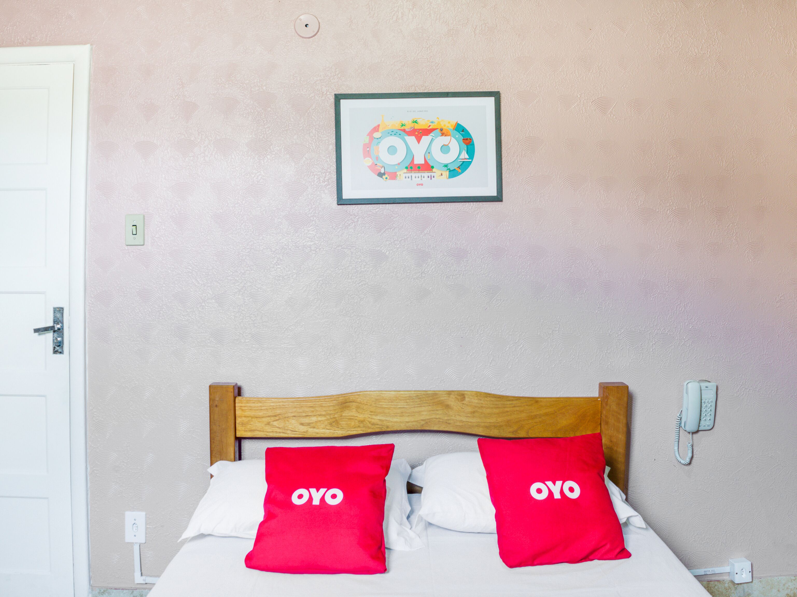 Quarto com cabeceira de madeira, duas almofadas vermelhas com a palavra 'OYO' e um quadro na parede, além de uma porta branca.