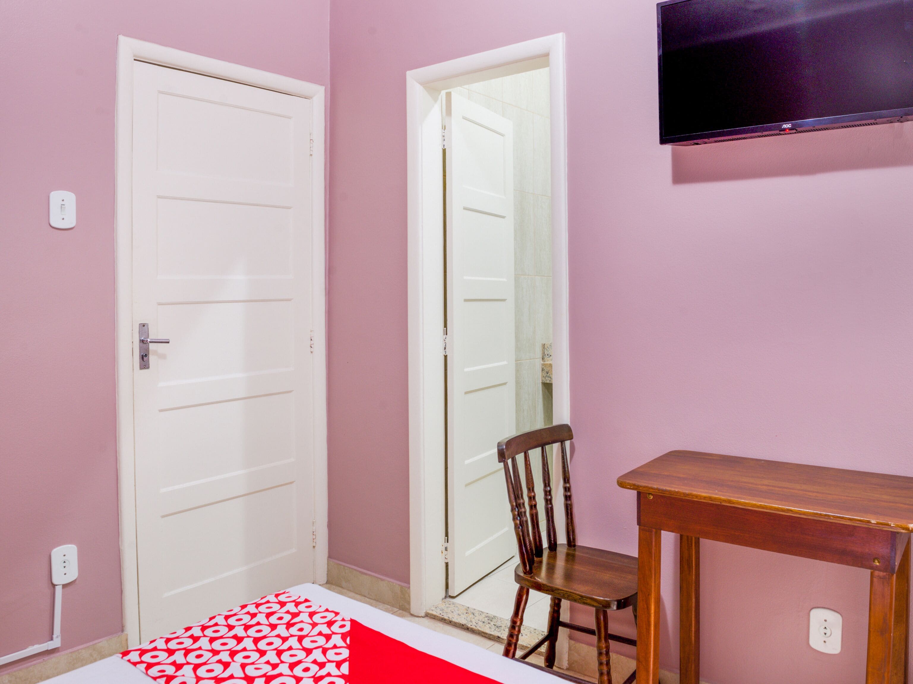 Una habitación con dos puertas blancas, una silla de madera, una pequeña mesa de madera y un televisor montado en la pared.