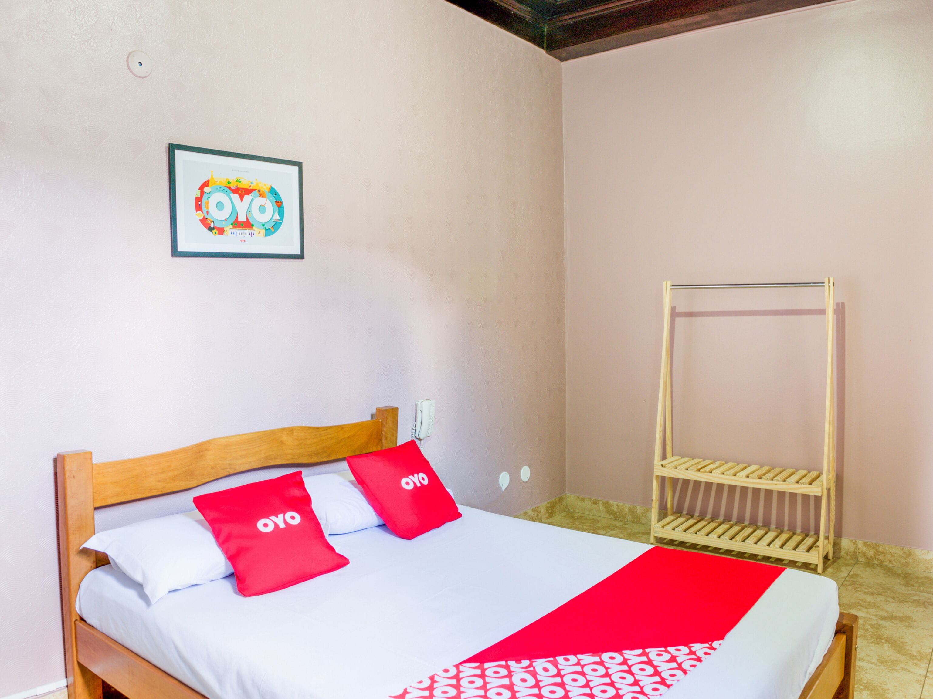 Una cama individual con estructura de madera, dos almohadas rojas, una colcha blanca y una manta roja. Hay una estantería de madera contra la pared y un cuadro enmarcado sobre la cama.