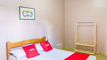 Una cama individual con estructura de madera, dos almohadas rojas, una colcha blanca y una manta roja. Hay una estantería de madera contra la pared y un cuadro enmarcado sobre la cama.