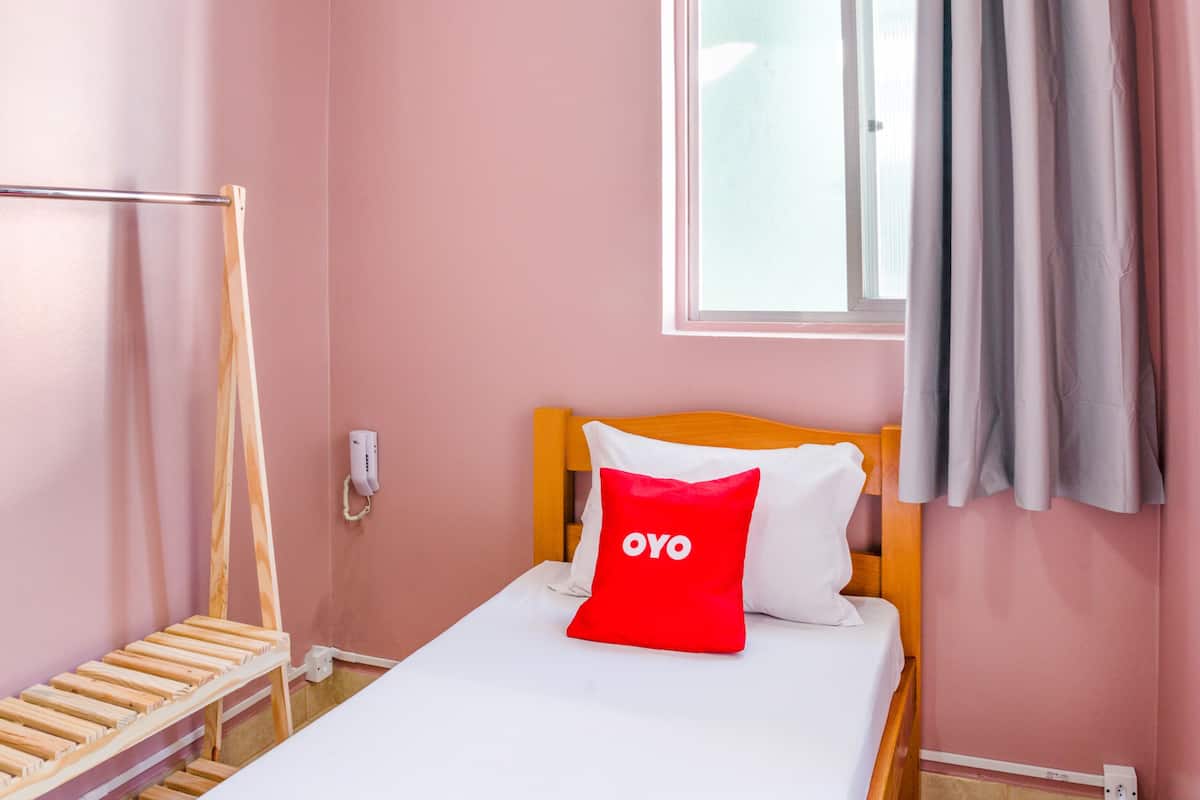 Ein Einzelbett mit Holz-Kopfstück, einem roten Kissen mit dem Aufdruck "OYO", einem weißen Kissen, einer Holzleiter und einem mit Vorhängen versehenen Fenster.