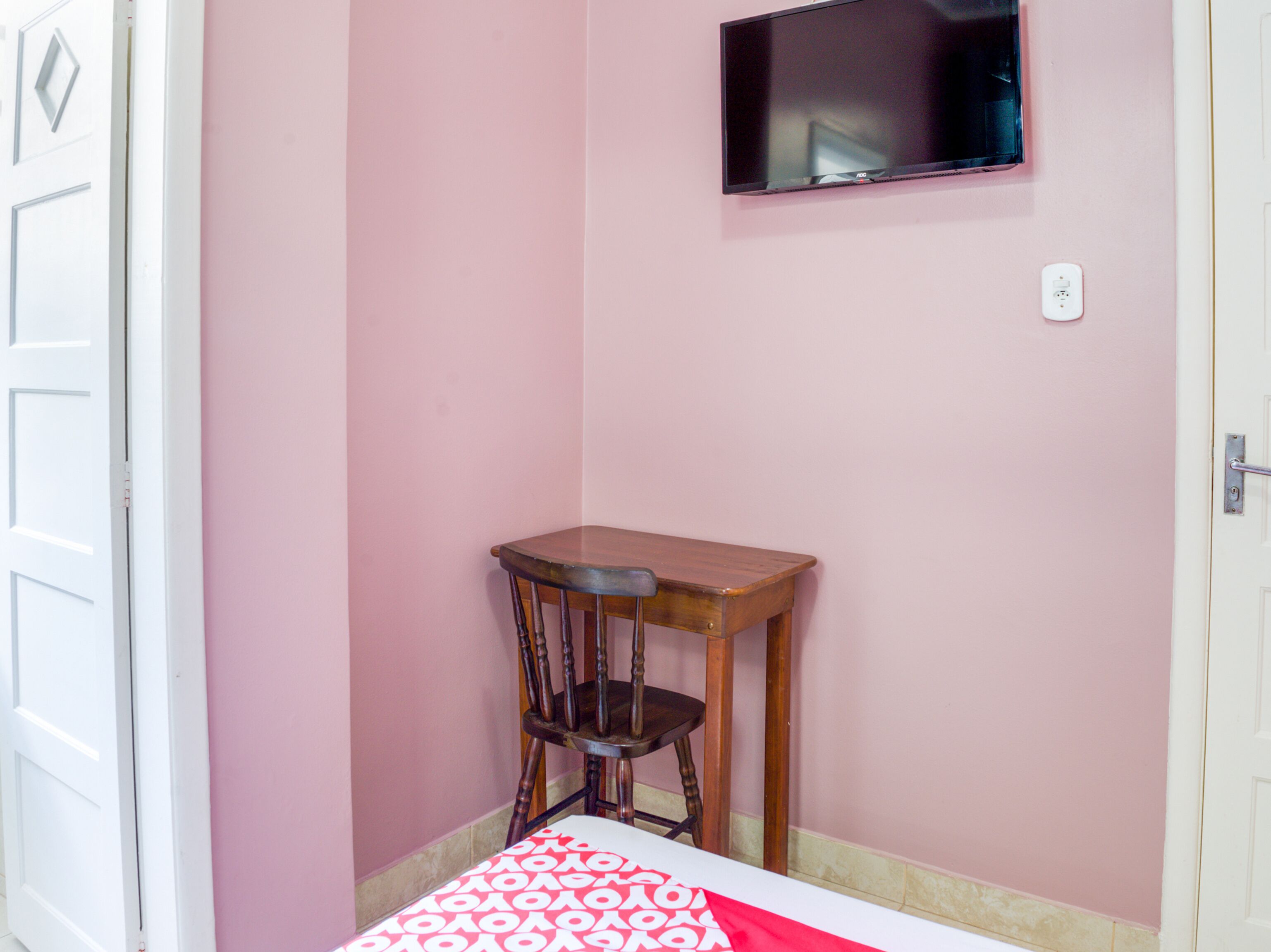 Quarto com cama, cadeira de madeira, mesa pequena e televisão fixada na parede.