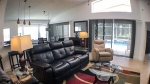 Un salon moderne avec un canapé en cuir noir, un fauteuil inclinable beige et une table basse en verre. On y trouve deux lampes, une grande fenêtre et une vue sur une piscine à l’extérieur.