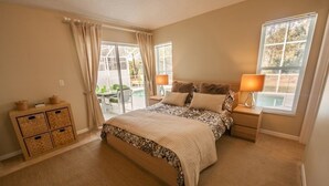 A bedroom with a large bed, a nightstand, a lamp, a window with blinds, and a sliding glass door leading to an outdoor area.