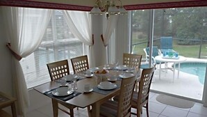 Een eetruimte met een tafel gedekt voor vier personen, grote ramen met witte gordijnen, en uitzicht op een zwembad en tuinmeubilair.
