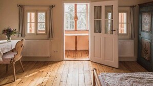 A room with wooden flooring, a dining table with chairs, a bed with a patterned bedspread, and a large window with curtains. - Stoantaler Höfl (Innsbruck)