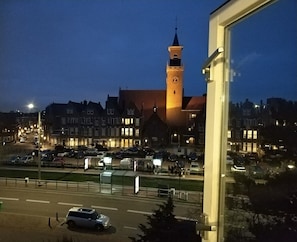 Une place de ville dominée par une tour imposante, entourée de bâtiments dont les fenêtres sont illuminées.