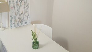 Une table blanche ornée d’une œuvre d’art à motifs de cactus, un petit vase vert contenant une plante et une chaise blanche.