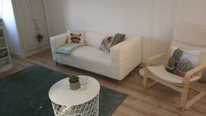 Un salon avec un canapé blanc, une table ronde blanche ornée d’une plante et de verres, une chaise en bois avec un cactus, un lampadaire et un tapis vert.