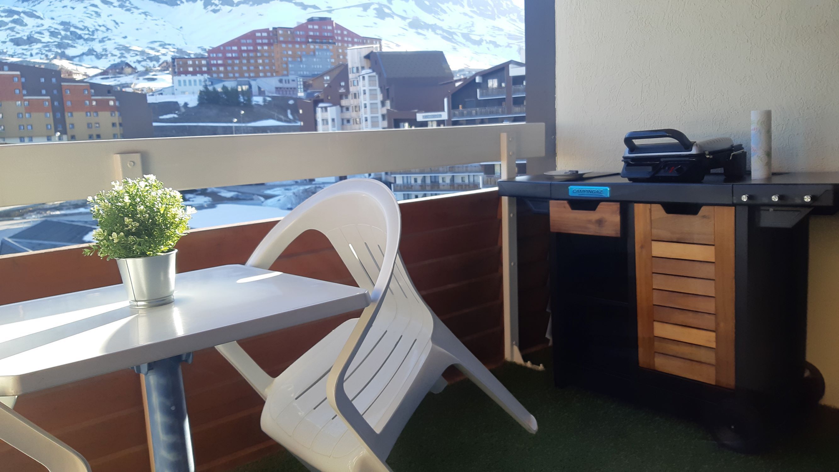 A balcony with a white plastic chair, a small potted plant, and a table. A desk with a printer is visible in the background.