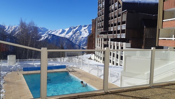 Una piscina con vista a montañas nevadas, rodeada por una cerca blanca y un edificio con balcones.