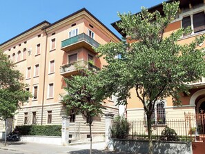 A multi-story building with balconies, a paved street, and a tree in the foreground.