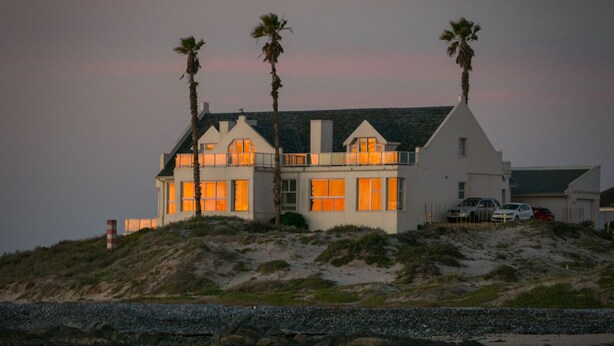 Una casa costera con un balcón destacado, ubicada en una colina arenosa con vistas al mar.