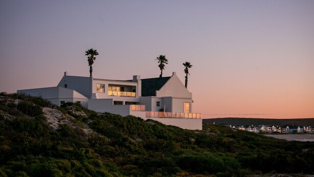 Una casa moderna frente al mar, con techo plano, ventanas amplias y dos palmeras.