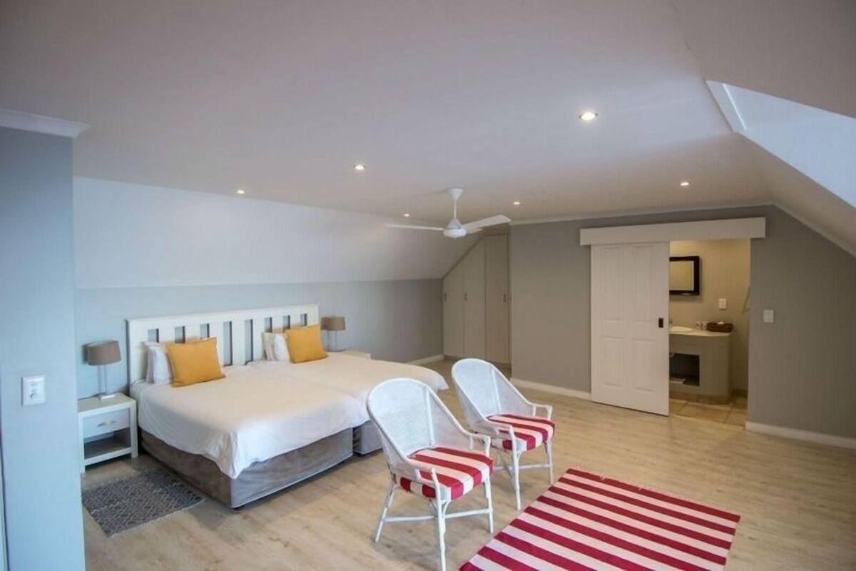 Quarto com duas camas, cadeiras brancas, tapete listrado em vermelho e branco, e um ventilador de teto.