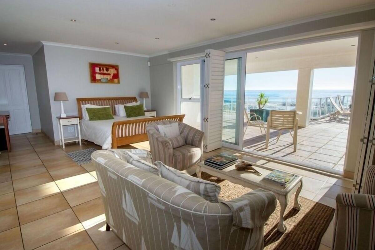 Uma sala de estar espaçosa com um sofá grande, uma cama com almofadas verdes e uma varanda com vista para o oceano.