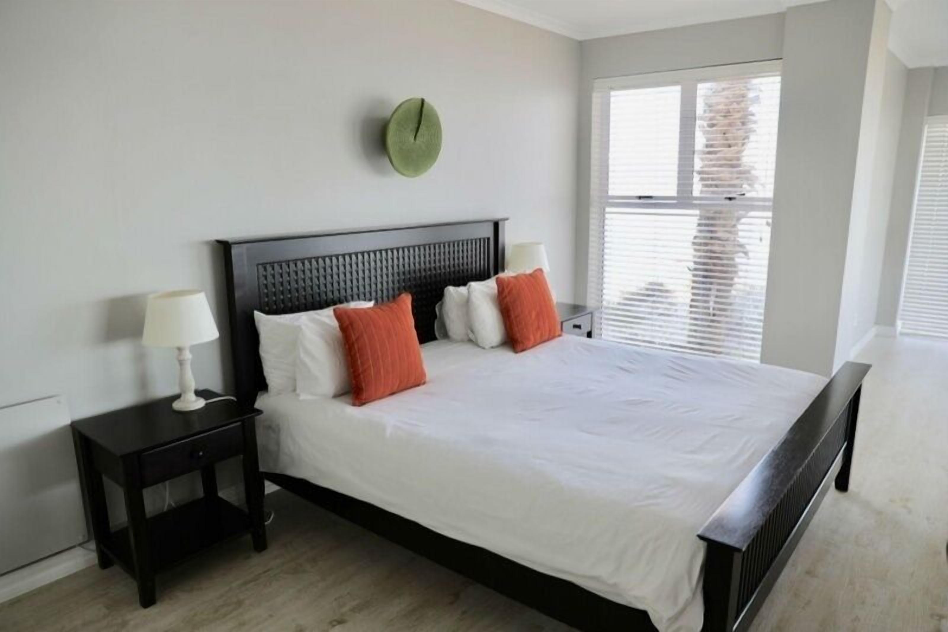 Une chambre à coucher avec un grand lit, deux tables de chevet, une horloge verte au mur et une fenêtre avec des stores.