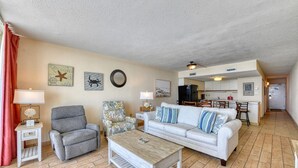 Ein Wohnzimmer mit einem grauen Sessel, einem blumigen Sessel, einem weißen Sofa mit blauen Kissen, einem Holztisch und einer Küche im Hintergrund.