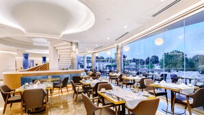 Un restaurant moderne doté d’un bar central, de nombreuses tables et de grandes fenêtres offrant une vue sur l’extérieur.