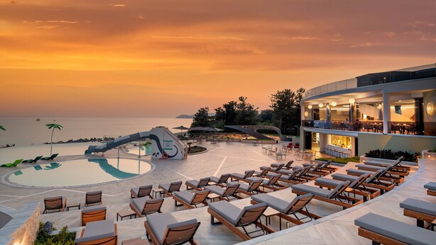 Un espace piscine aménagé dans un complexe hôtelier, avec des chaises longues et un bâtiment moderne abritant un bar.