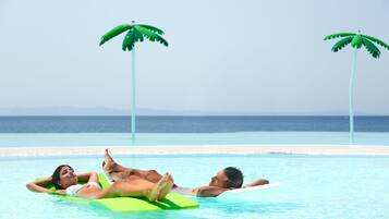 A pool with two people lying on floatation devices, surrounded by a clear blue sky and a calm sea in the background.